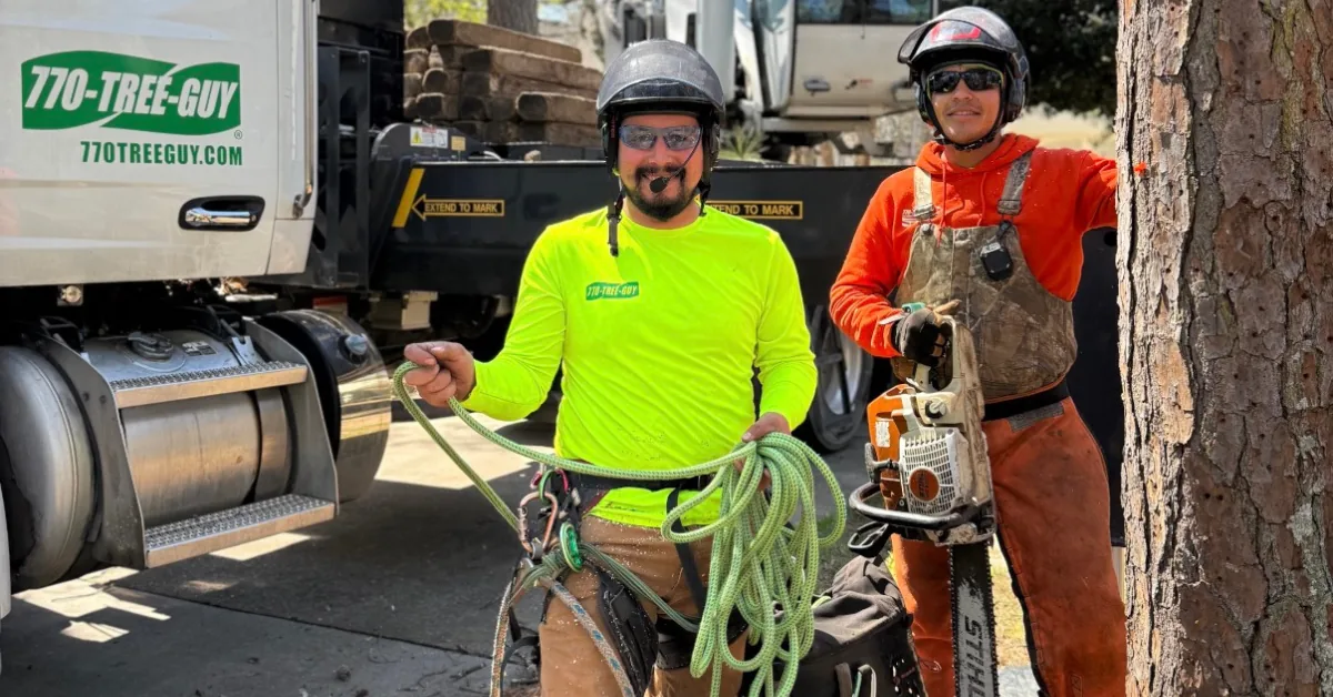 Two tree crew members in safety helmets and chaps pose with a chainsaw and climbing rope by the truck.